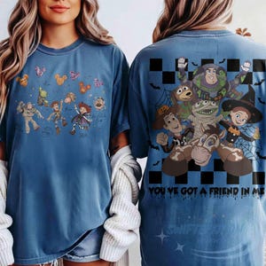 May include: A blue t-shirt featuring Toy Story characters in Halloween costumes. The front shows small character illustrations with Mickey-shaped balloons. The back displays a large graphic of Buzz Lightyear, Woody, and friends with a black and white checkerboard pattern, spiderwebs, and the text "YOU'VE GOT A FRIEND IN ME."