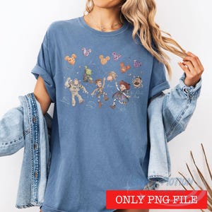 May include: A blue short-sleeved t-shirt featuring a graphic print of various animated characters including a space ranger, a cowboy, and a cowgirl, accompanied by floating Mickey Mouse-shaped balloons and handwritten-style text labels.