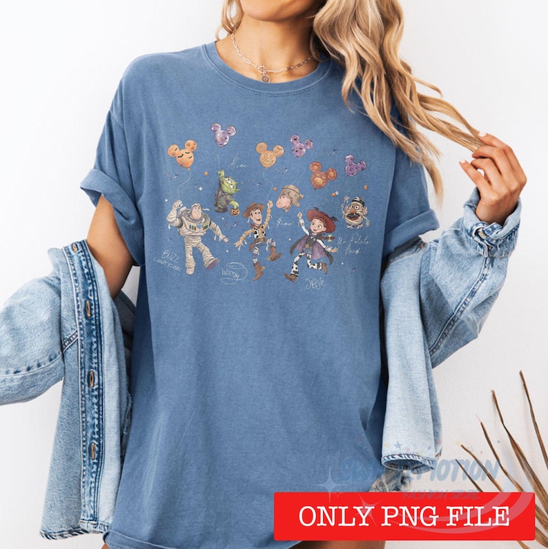 May include: A blue short-sleeved t-shirt featuring a graphic print of various animated characters including a space ranger, a cowboy, and a cowgirl, accompanied by floating Mickey Mouse-shaped balloons and handwritten-style text labels.