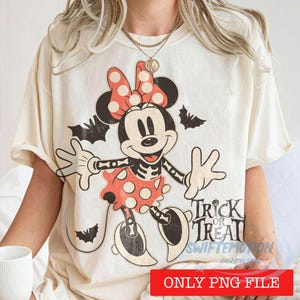 May include: Cream-colored t-shirt featuring a Halloween-themed Minnie Mouse design. Minnie is depicted with a skeleton body, wearing a red polka-dot bow and dress. The shirt includes the text "Trick or Treat" and bat silhouettes.