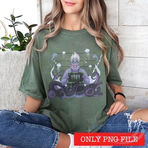 May include: An olive green t-shirt featuring a graphic of a purple sea witch character with tentacles, holding a black sign with the text 1989 in green. The background includes a mugshot height chart, jellyfish, and eels in a stylized illustration.