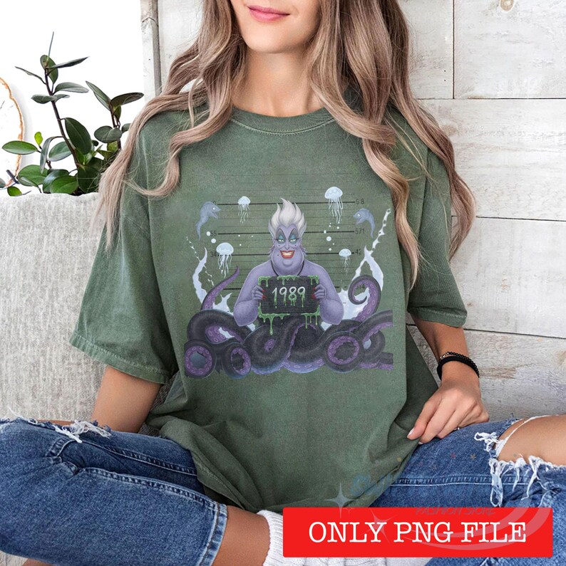 May include: An olive green t-shirt featuring a graphic of a purple sea witch character with tentacles, holding a black sign with the text 1989 in green. The background includes a mugshot height chart, jellyfish, and eels in a stylized illustration.