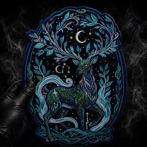 Puede incluir: Un parche bordado de forma ovalada que muestra un ciervo con grandes astas hechas de hojas. El diseño incluye una luna creciente, estrellas y patrones de remolinos azules y verdes sobre un fondo negro, con un intrincado trabajo de hilo.