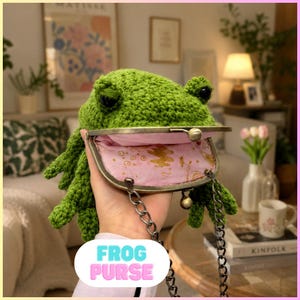 May include: A green crocheted frog-shaped purse with a metal clasp and chain strap. The interior is lined with pink fabric. The purse has a frog face design and the text "FROG PURSE" in a bubble font.
