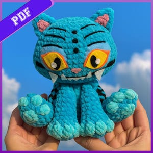 Puede incluir: Un juguete de peluche de gato azul con acabado de hilo texturizado, con grandes ojos amarillos y naranjas, marcas negras en la frente y colmillos de fieltro blanco. La criatura tiene una pequeña nariz rosa y almohadillas azul claro. Banner PDF.