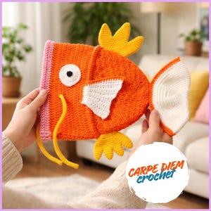 May include: A crocheted fish-shaped item in bright orange with a white eye, white side fin, yellow top and bottom fins, and two long yellow whiskers. The front edge is light pink. A white circular logo features the text CARPE DIEM crochet.