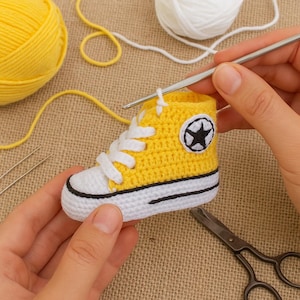Op de afbeelding: Een handgemaakte gele en witte gehaakte babysneaker met een zwart sterembleem. De miniatuurschoen heeft witte veters en een zwarte streep langs de zool. Haaknaalden, garen en schaar staan op de achtergrond.