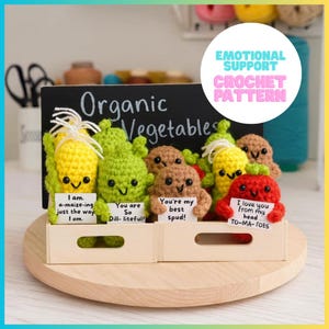 May include: A collection of small, colorful crocheted vegetables including corn, a pickle, a potato, and a tomato, each holding a white sign with punny text. They are displayed in two light-colored wooden crates in front of a chalkboard sign.