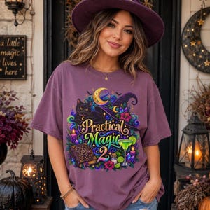 May include: A mauve short-sleeved t-shirt featuring a graphic print of a black cat, a spellbook, a witch hat, and a cocktail glass with the text "Practical Magic 2". The shirt is worn with a purple witch hat adorned with a gold star accent.