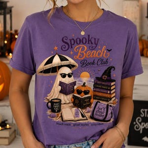 May include: A purple t-shirt featuring a graphic print with the text Spooky Beach Book Club. The design shows a ghost wearing sunglasses under a beach umbrella, a pumpkin reading a book, a stack of books, and a tote bag labeled hot ghoul reader.