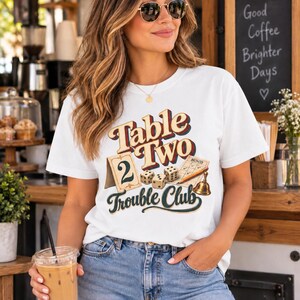 May include: A white short-sleeve t-shirt featuring a graphic print with the text Table Two Trouble Club. The design includes a table number card with the number 2, three white dice, a Bunco score sheet, a pencil, and a small brass bell.