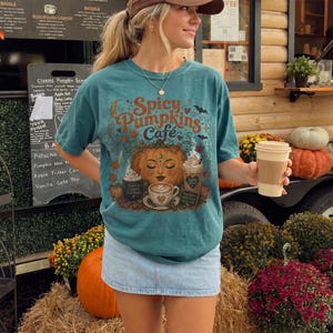 May include: A teal short-sleeve t-shirt featuring a graphic print with the text Spicy Pumpkins Cafe, a central pumpkin face, coffee cups, and autumn motifs. The shirt is paired with a light blue denim skirt and a brown baseball cap.
