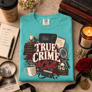 May include: A teal t-shirt featuring a graphic print with the text True Crime & Chill, a television, a stack of books, a magnifying glass, a skull, a cassette tape, and a coffee mug with the text Good People Binge Bad Things.