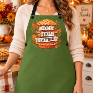 May include: An olive green apron featuring a central graphic of three stacked pies surrounded by autumn leaves and berries. The text "PIE FIXES EVERYTHING" is printed on three white banners layered across the pies. The apron has black straps.