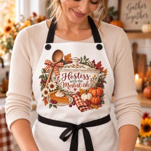 May include: A white apron with black straps and buttons featuring a graphic of a casserole dish, a wooden spoon, a slice of pumpkin pie, and autumn foliage. The text reads Hostess with the Mostest-ish in a decorative red script font.