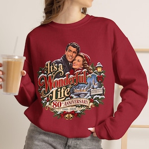 May include: A dark red crewneck sweatshirt featuring a graphic print of the movie It's a Wonderful Life. The design includes a couple, a snowy town scene, a street lamp, and text reading It's a Wonderful Life 80th Anniversary 1946-2026.