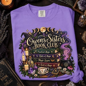 May include: A light purple t-shirt featuring a graphic print with the text Owens Sisters Book Club. The design includes a stack of four books, a witch hat, a steaming cauldron, candles, a teacup, crystals, and moon phases in a mystical style.