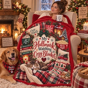 May include: A red fleece blanket featuring a festive holiday design with the text My Hallmark Watching Cozy Blanket, surrounded by illustrations of hot cocoa, a sleeping cat, a red truck, pine trees, and stockings, with a plaid pattern border.