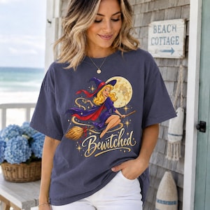 May include: A dark gray short-sleeve t-shirt featuring a graphic of a blonde witch in a purple dress and hat flying on a broomstick in front of a full moon. The word Bewitched is printed in a gold cursive script below the illustration.