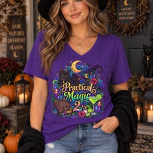 May include: A purple V-neck t-shirt featuring a graphic print of a black cat, a crescent moon, a witch hat, a spellbook, a cocktail glass, and candles. The text reads Practical Magic 2 in colorful, stylized lettering surrounded by swirling patterns.
