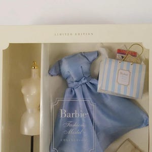 以下が含まれることがあります： クリーム色の箱に入った限定版バービーファッションモデルコレクションセット。ウエストに小さなリボンが付いたライトブルーのドレス、黒いスタンドに乗った白いマネキン、バービーのロゴが入った白と青のストライプのショッピングバッグが含まれています。