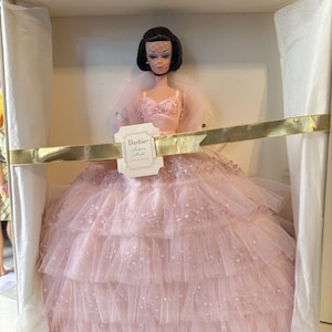 May include: A vintage Barbie doll in a box. The doll wears a pink, layered tulle gown with sequin accents and a matching bodice. The doll has dark hair and is presented in a white box with a gold ribbon.