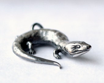 May include: A silver-coloured lizard pendant. The reptile is curled, with visible legs, a tail, and a loop for a chain. The pendant is likely metal and set against a white background. The lizard measures approximately 5 cm in length.