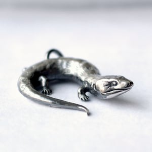 May include: A detailed, silver-toned lizard pendant. The reptile is depicted in a curled pose, with visible legs, a tail, and a loop for a chain. The pendant is likely made of metal and is set against a white background. The lizard measures approximately 2 inches in length.