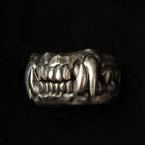 May include: A silver ring with a detailed design of a set of teeth, resembling a predator's jaw.