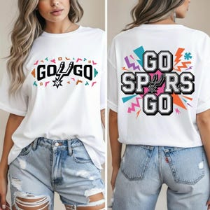 Może przedstawiać: Biała koszulka z napisem "GO SPURS GO" z przodu i z tyłu. Przedni wzór zawiera logo drużyny i słowa "GO GO" w kolorowym wzorze. Tylny wzór zawiera słowa "GO SPURS GO" z kolorowym tłem.