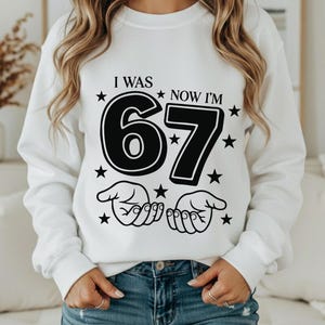 以下が含まれることがあります： 黒い文字で「I WAS 67 NOW I'M」と書かれた白いスウェットシャツ。数字の67は太字で、星と手のイラストがデザインされています。青いジーンズを履いています。