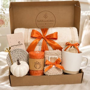 May include: A cardboard gift box from SoftResetGiftCo containing a cream plaid blanket tied with an orange ribbon, a white mug, a small burlap bag, a white ceramic pumpkin, a textured coaster, a gold spoon, and an orange candle with a pumpkin drawing.