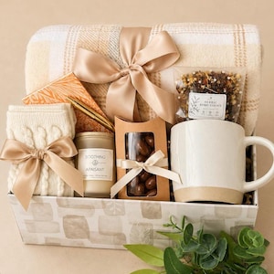 May include: A gift basket containing a beige plaid throw blanket with a satin ribbon, a cream cable-knit pair of socks, a gold pen, a candle labeled Soothing Apaisant, a box of chocolates, a bag of herbal berry essence, and a two-tone ceramic mug.
