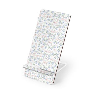 Pode incluir: Um suporte de telefone branco com um padrão floral de flores rosa e azuis. O suporte tem uma prateleira de plástico transparente para segurar um telefone e uma base branca. O padrão floral cobre a frente.