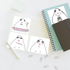 May include: A spiral-bound notebook with a light blue cover featuring four distinct white stickers. Each sticker displays a hand-drawn, white, blob-like character with a pink nose, shown in various poses including wearing a pink tutu, holding a star wand, looking grumpy, and smiling with rosy cheeks.