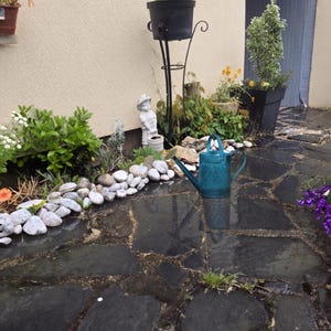 Peut inclure: Un arrosoir en métal bleu sarcelle avec un motif en losange texturé est posé sur une terrasse en pierre sombre. À proximité, une rangée de galets blancs borde un massif contenant des plantes vertes, une petite statue blanche et un support.
