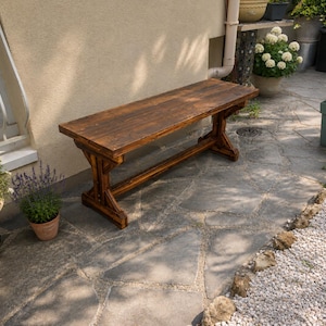 Può includere: Una panca rettangolare in legno con finitura tinta marrone scuro. La panca presenta una seduta piatta sostenuta da due robuste gambe a cavalletto collegate da una barra trasversale orizzontale, che fornisce una base stabile per questo sedile.