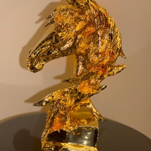 May include: A gold-colored horse head sculpture featuring a highly textured, reflective surface. The piece depicts the head and neck of a horse with a flowing mane, mounted on a solid, smooth, rectangular base that catches the light.
