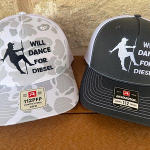 May include: Two trucker hats with white mesh backs. The left hat features a light gray camouflage pattern with a black silhouette graphic and the text "WILL DANCE FOR DIESEL." The right hat is solid dark gray with a white silhouette and the same text.
