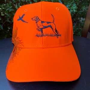 May include: Orange baseball cap with a black embroidered design of a dog pointing at a pheasant. The cap has a brown and gold camouflage pattern on the side.