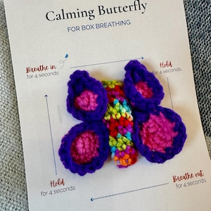 May include: A crochet butterfly in shades of purple, pink, and rainbow colors, resting on a white card. The card reads "Calming Butterfly FOR BOX BREATHING" with instructions: "Breathe in for 4 seconds," "Hold for 4 seconds," and "Breathe out for 4 seconds."