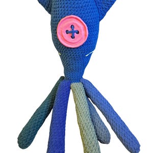 May include: A handmade crochet plush toy shaped like a squid with a dark blue body, a large pink button eye, and multiple dangling tentacles in shades of blue and muted green. The textured yarn construction features a pointed head and long limbs.