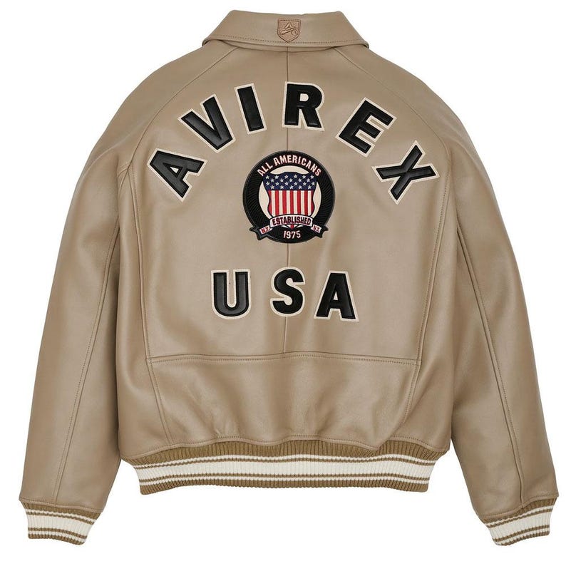 May include: A tan leather bomber jacket featuring a ribbed collar, cuffs, and hem with white stripes. The back displays the black embroidered text AVIREX and USA, along with a circular patch featuring a flag design and the text ALL AMERICANS.