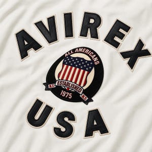 Op de afbeelding: Een gebroken wit kledingstuk met de zwarte lederlook applicatietekst AVIREX en USA. In het midden bevindt zich een ronde badge met een schildafbeelding, sterren, strepen en de tekst ALL AMERICANS ESTABLISHED 1975 N.Y. in een zwart en rood kleurenschema.
