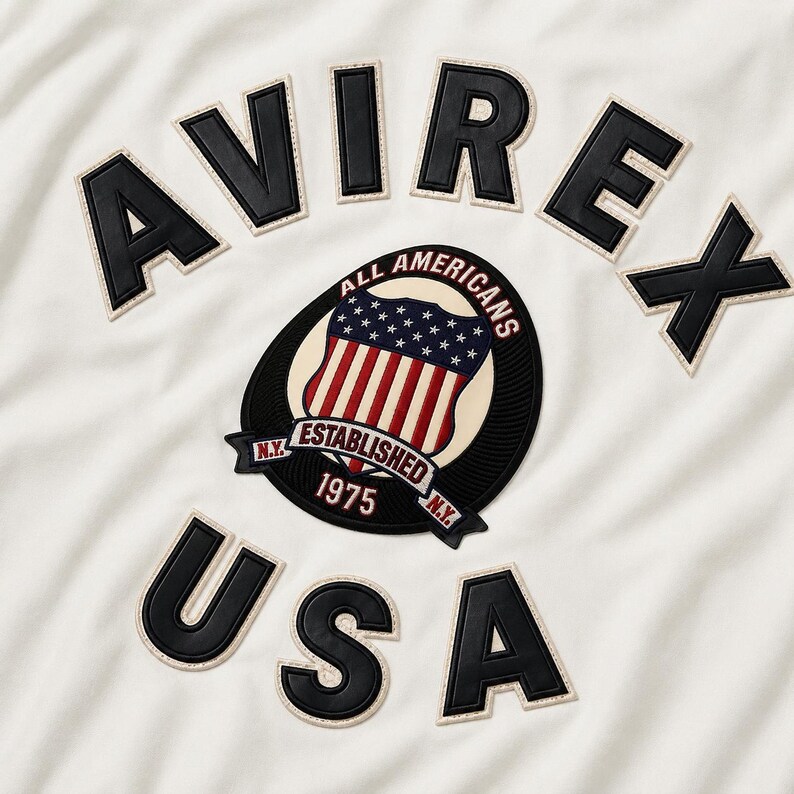 Op de afbeelding: Een gebroken wit kledingstuk met de zwarte lederlook applicatietekst AVIREX en USA. In het midden bevindt zich een ronde badge met een schildafbeelding, sterren, strepen en de tekst ALL AMERICANS ESTABLISHED 1975 N.Y. in een zwart en rood kleurenschema.