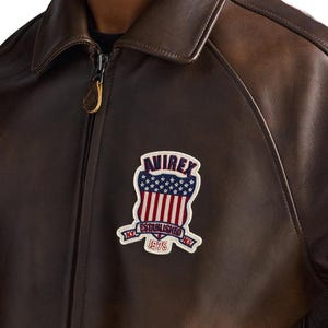 May include: A dark brown leather jacket featuring a full-length front zipper with a branded pull tab. On the chest, a patch displays the text AVIREX, ESTABLISHED, 1975, and N.Y. with a graphic of a shield containing stars and vertical stripes.
