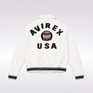 Op de afbeelding: Een wit jack met de zwarte tekst AVIREX USA op de achterkant. In het midden tussen de woorden staat een rond embleem met een rood, wit en blauw Amerikaanse vlag-icoon, de tekst ALL AMERICANS en het jaartal 1975. De boorden en de zoom zijn zwart-wit gestreept.