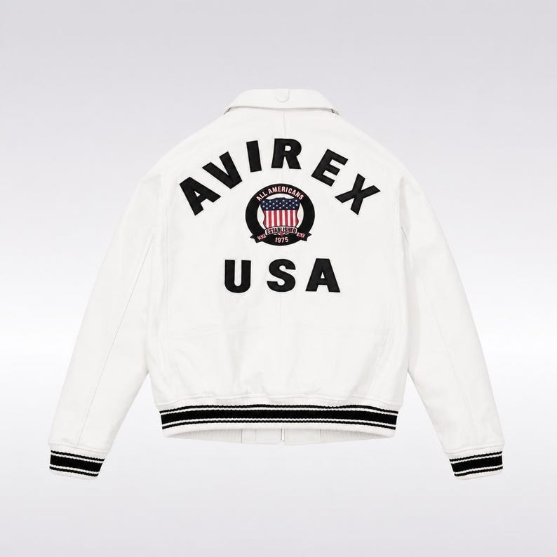Op de afbeelding: Een wit jack met de zwarte tekst AVIREX USA op de achterkant. In het midden tussen de woorden staat een rond embleem met een rood, wit en blauw Amerikaanse vlag-icoon, de tekst ALL AMERICANS en het jaartal 1975. De boorden en de zoom zijn zwart-wit gestreept.