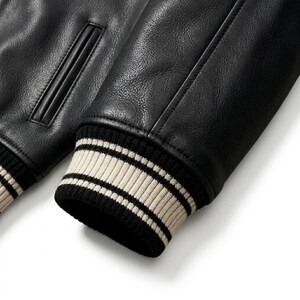 May include: A black leather jacket sleeve featuring a textured surface and a ribbed knit cuff. The cuff is designed with alternating horizontal stripes in cream and black colors. A vertical welt pocket is visible on the upper section of the jacket.