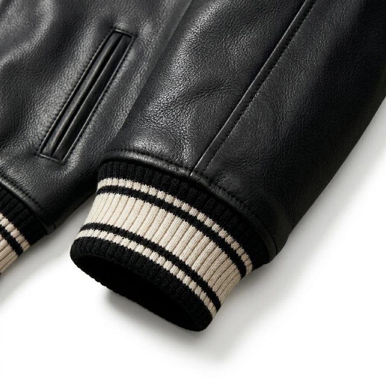 May include: A black leather jacket sleeve featuring a textured surface and a ribbed knit cuff. The cuff is designed with alternating horizontal stripes in cream and black colors. A vertical welt pocket is visible on the upper section of the jacket.